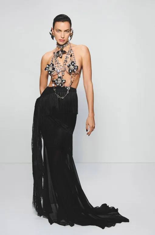 Ирина Шейк в длинном чёрном платье Blumarine с открытым декольте и эффектным декоративным элементом на груди, студийный свет и минимализм фона.