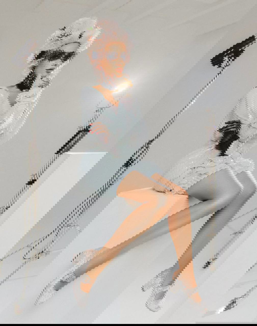 Маш Милаш в нежном пастельном платье и украшениях рококо, сидит на белом кубе; кадр подчёркивает складки ткани и детальную обувь внизу.