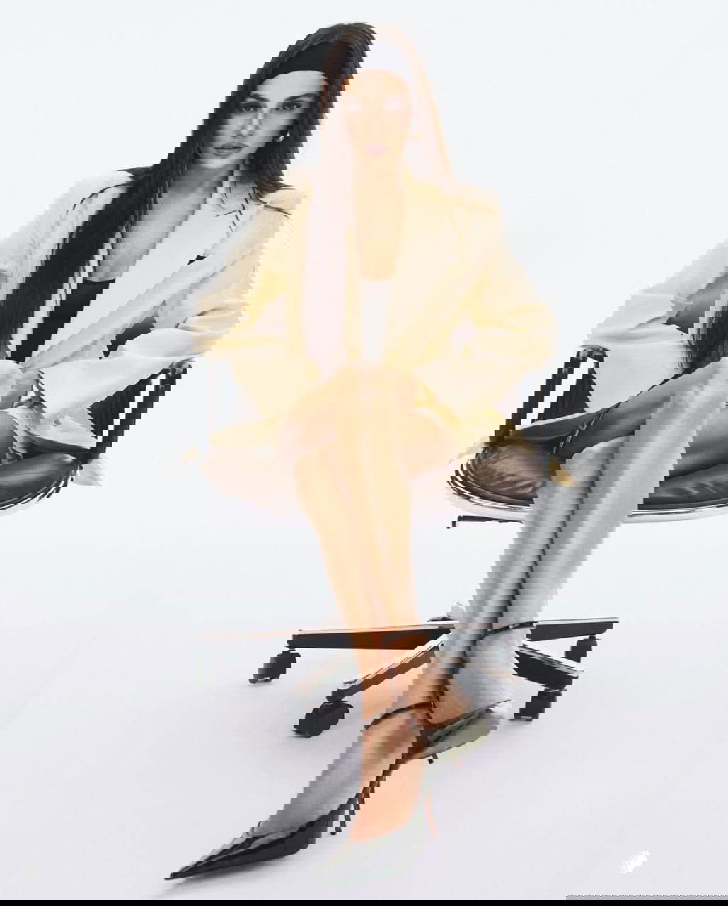 Женщина-актриса в светлом жакете в офисном кресле, строгий стиль и элегантная поза на белом фоне фотосессии.