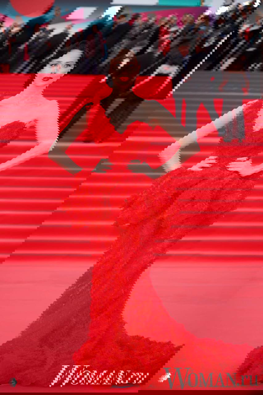 Женщина в пышном красном платье с объёмным декором позирует на красной дорожке 48-го ММКФ, выразительный макияж и постановочная фигура.