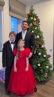 Мужчина в костюме с двумя детьми на фоне высокой украшенной ёлки — формальная семейная фотография в праздничной обстановке.