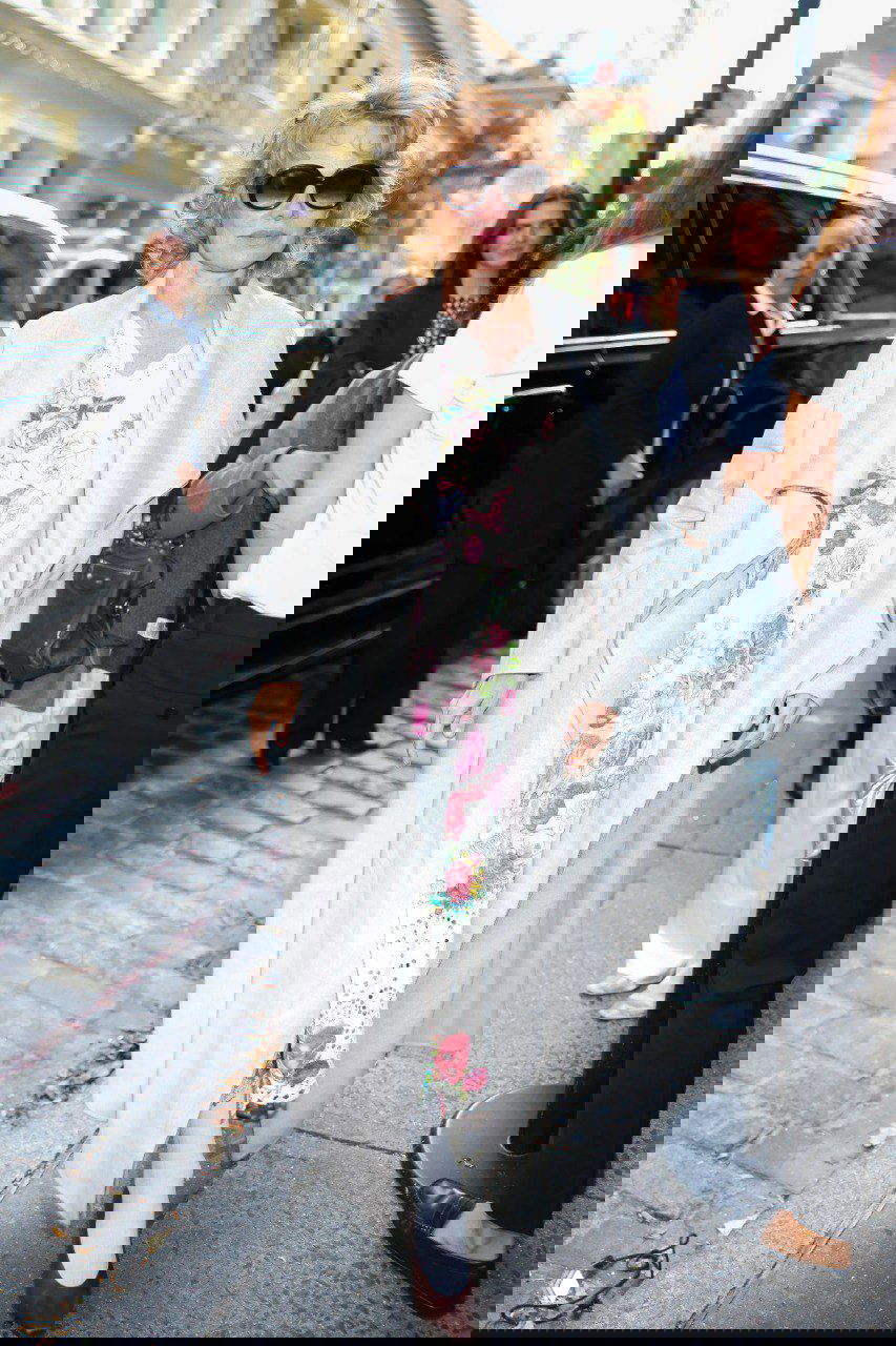 Памела Андерсон в пальто и платье в цветочный принт выходит на улицу у входа в pop-up магазин, образ в романтическом стиле с солнечными очками.