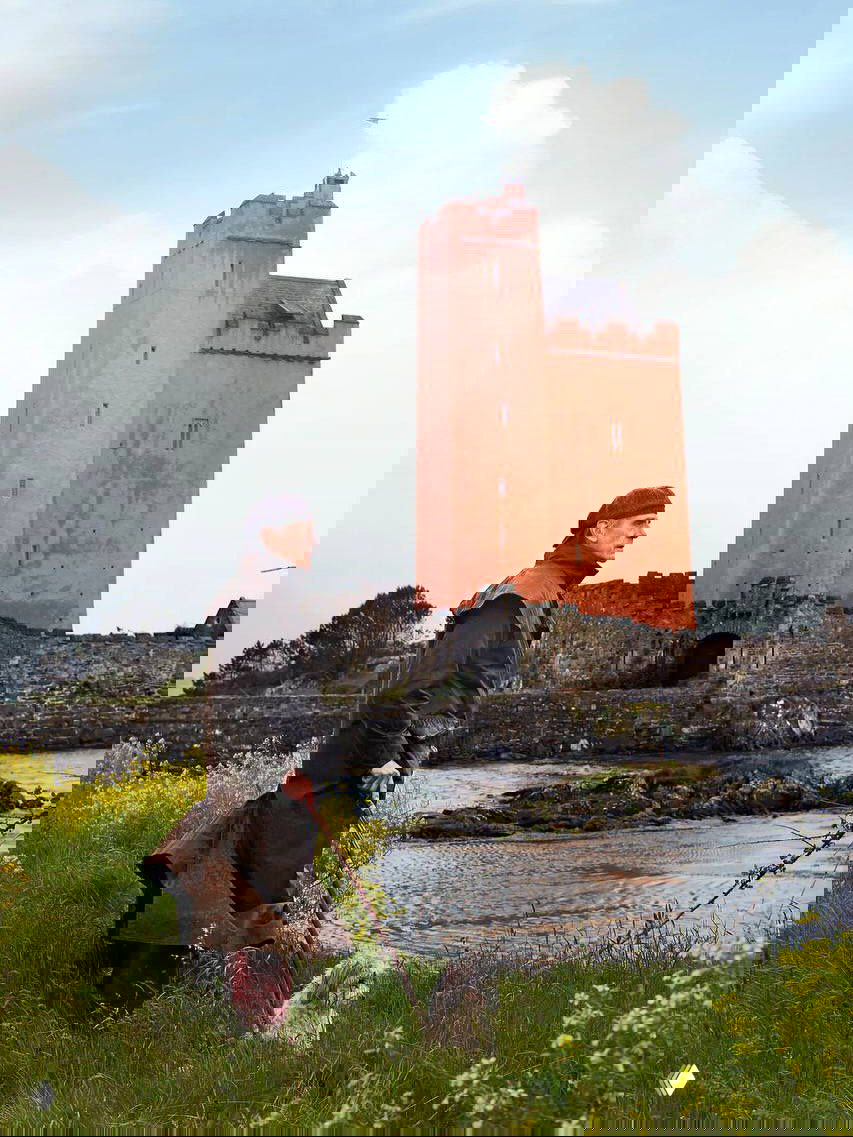 Внешний вид красочной башни замка Килко на фоне неба; на переднем плане Джереми Айронс стоит с прогулочной палкой и собакой у береговой линии.