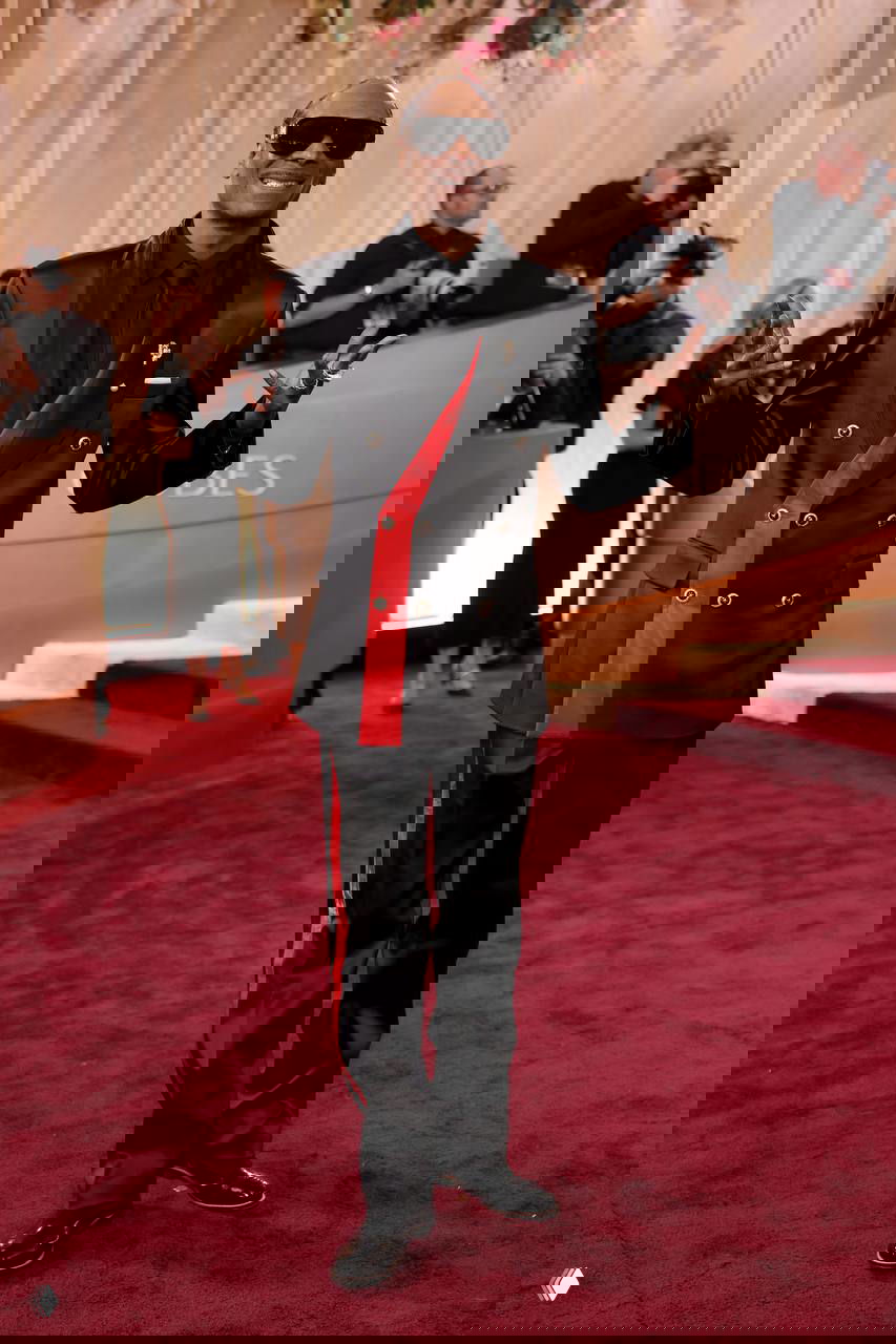 Мужчина в черном костюме с красной вставкой улыбается на красной дорожке «Золотого глобуса», вокруг него — фотографы и гости мероприятия.