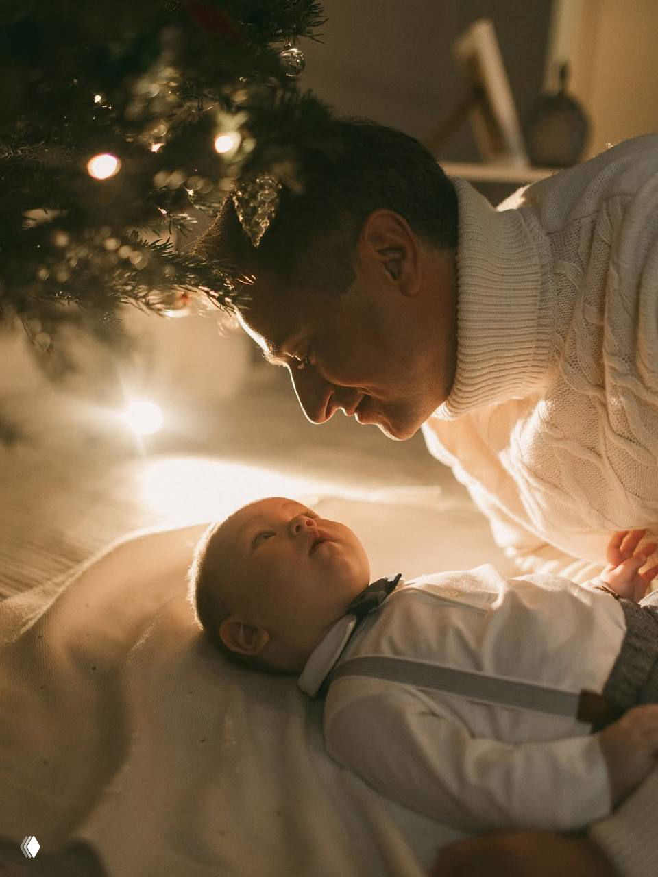 Мужчина наклоняется над малышом под ёлкой: нежный момент в тёплом боковом свете с акцентом на взгляде ребёнка.