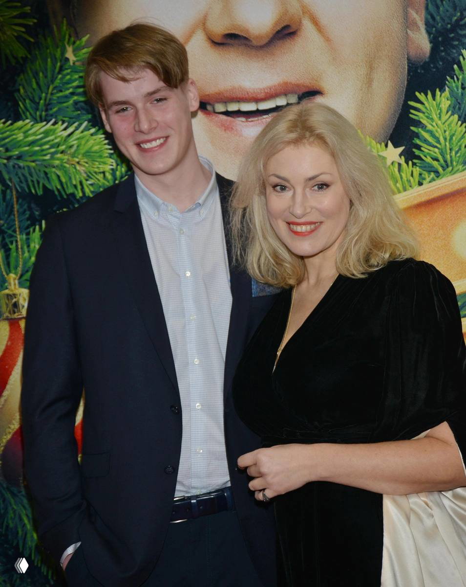 Крупный кадр: Мария Шукшина улыбается рядом с молодым мужчиной на фоне постера премьеры; видно лица и праздничный баннер позади них.