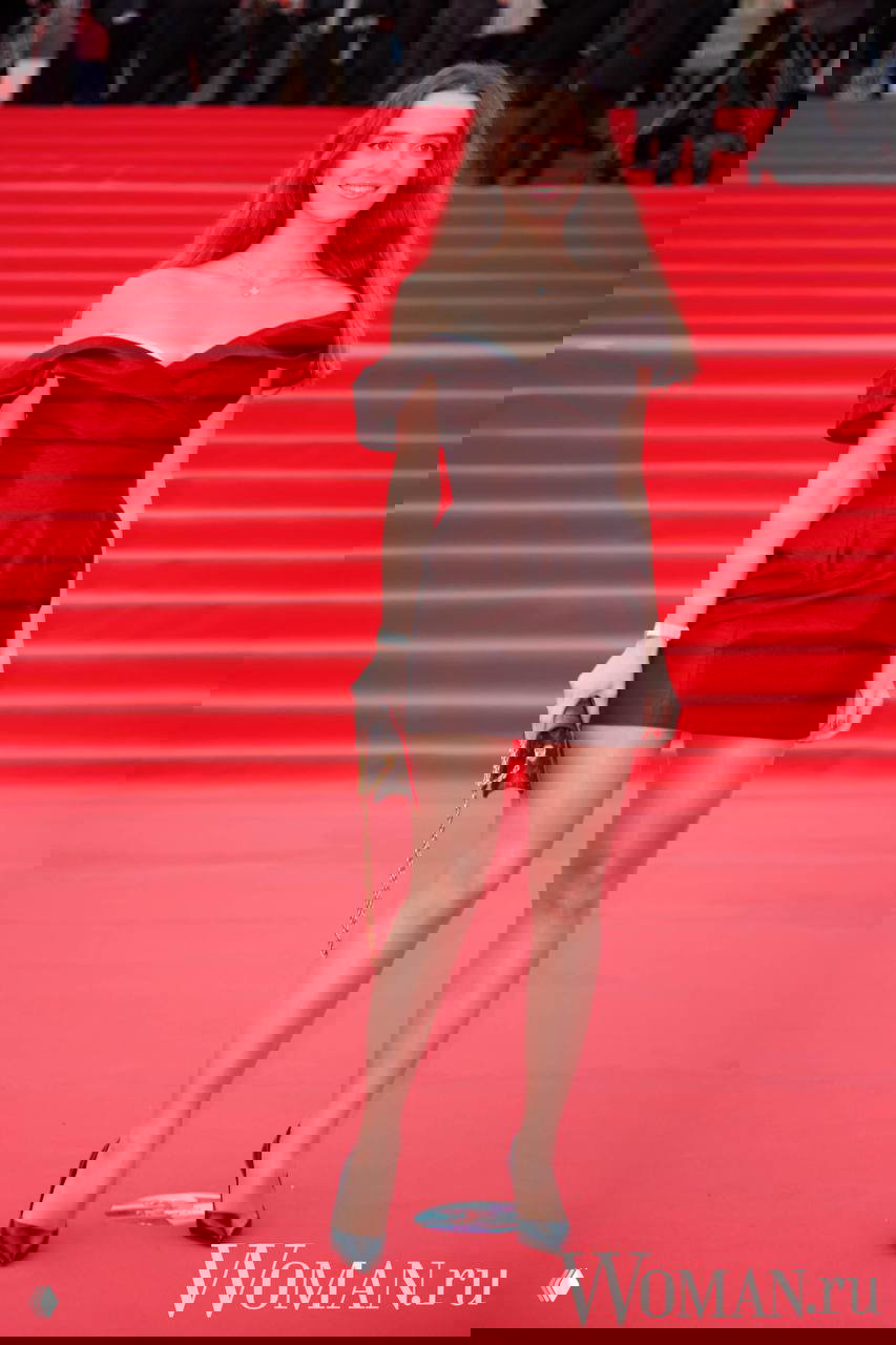 Молодая женщина в коротком тёмном платье и туфлях на каблуке стоит на красной дорожке 48-го ММКФ с небольшим клатчем, вокруг толпа и папарацци.