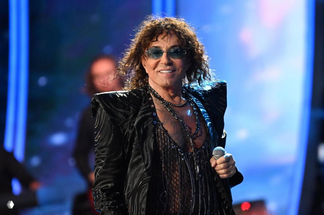 Мужчина‑исполнитель в тёмном сценическом костюме с украшениями держит микрофон на фоне яркого светового экрана, концертный кадр крупным планом.