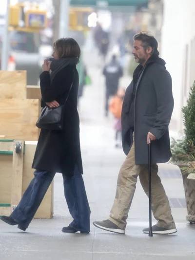 Уличный кадр: пара идёт по тротуару Манхэттена в пальто, мужчина держится за трость; на заднем плане прохожие и город.