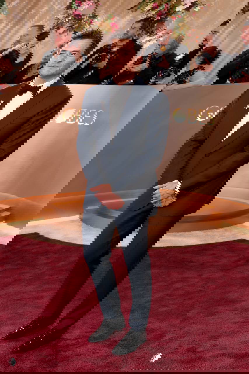 Мужчина в темном костюме с брошей позирует на красной дорожке «Золотого глобуса», вокруг видны фотографы и часть логотипа мероприятия.