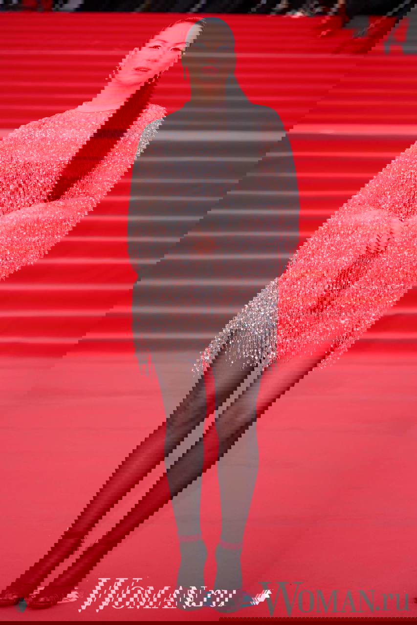 Женщина в блестящем платье с бахромой позирует фронтально на красной дорожке 48-го ММКФ, видна театральная лестница и фотографы на заднем плане.