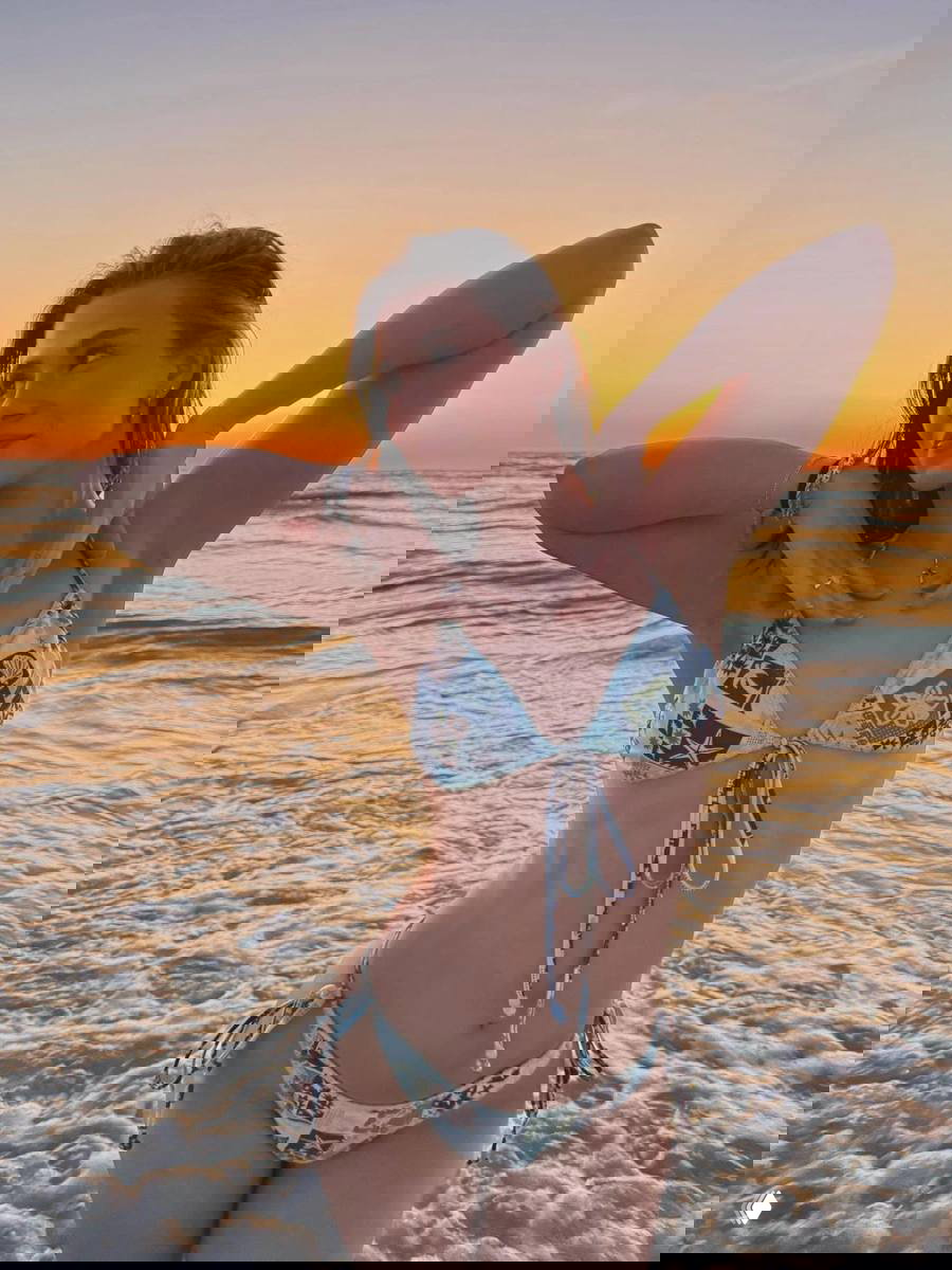 Девушка в бикини на берегу моря на фоне оранжевого заката; поза с поднятыми руками и тёплые оттенки неба над линией горизонта и волнами.