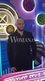 Полноразмерный кадр мужчины-музыканта в чёрном костюме на фоне сцены с логотипом премии и яркой декорацией, заметен водяной знак Woman.ru.