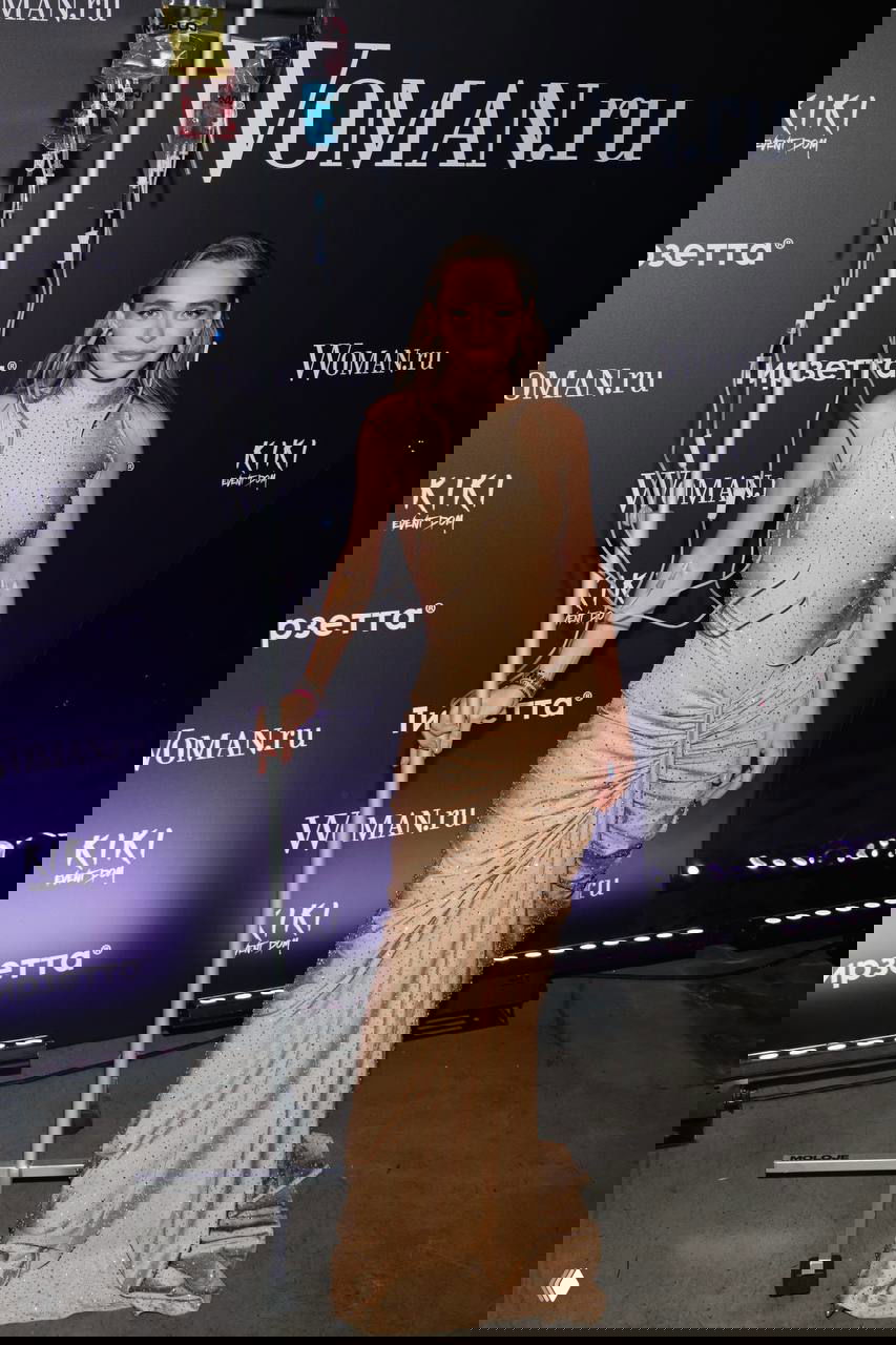 Гостья в бежевом облегающем платье позирует у брендволла; рядом декоративная стойка с инфузионными мешками как реквизит.