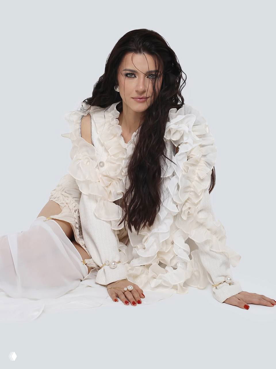 Женщина в белом рюшистом костюме сидит на светлом фоне, руки сложены, видно жемчужные украшения и декоративные элементы одежды