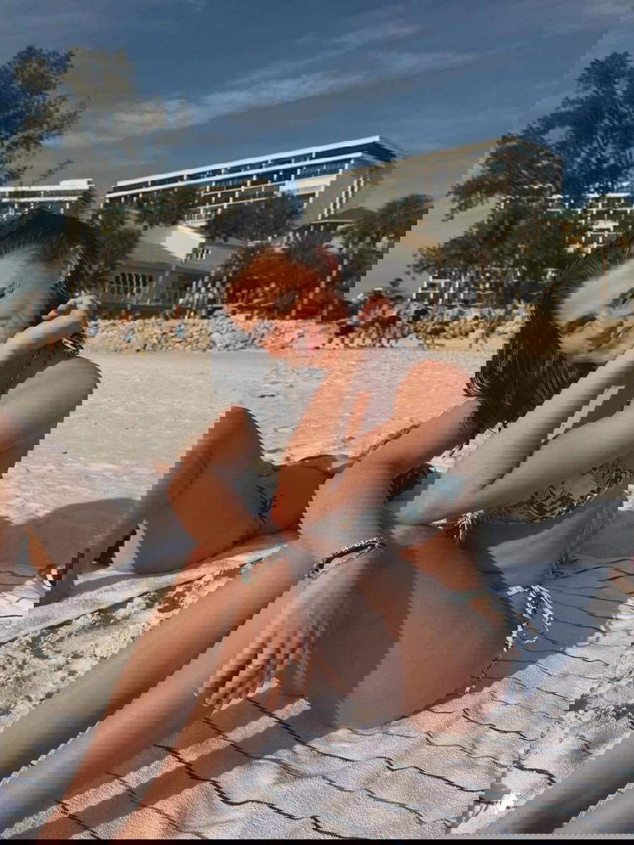 Молодая женщина в бикини сидит на песке у моря; солнечный свет, люди и гостиничные здания на заднем плане создают атмосферу пляжного отдыха.