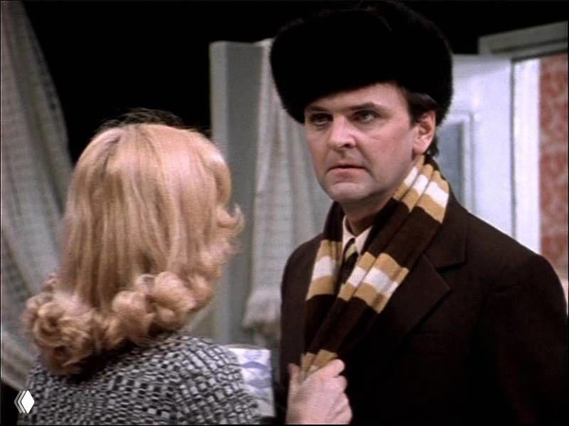 Кадр с мужчиной в тёплой шапке и шарфе, обращённым к женщине; зимняя одежда и интерьер подчёркивают новогоднюю атмосферу фильма.