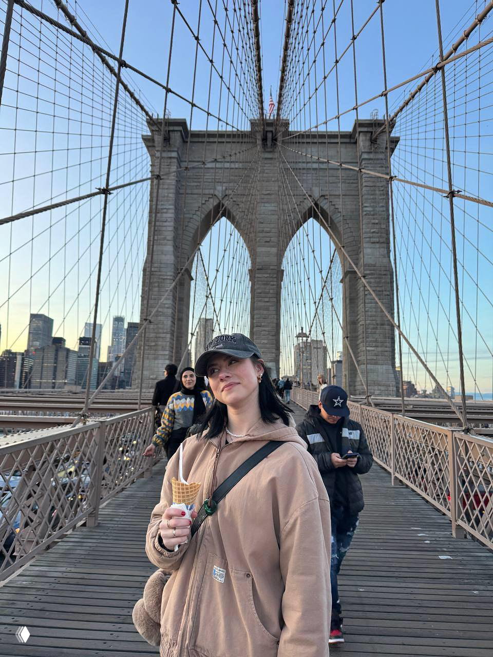 Женщина в худи и кепке идёт по пешеходной части большого арочного моста с рожком мороженого в руке; городской пейзаж и пешеходы вокруг.