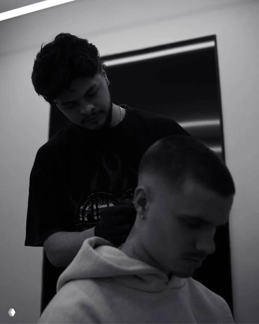 Чёрно‑белая фотография: мастер наносит татуировку на затылок молодого мужчины в худи, кадр показывает процесс и концентрацию работы.