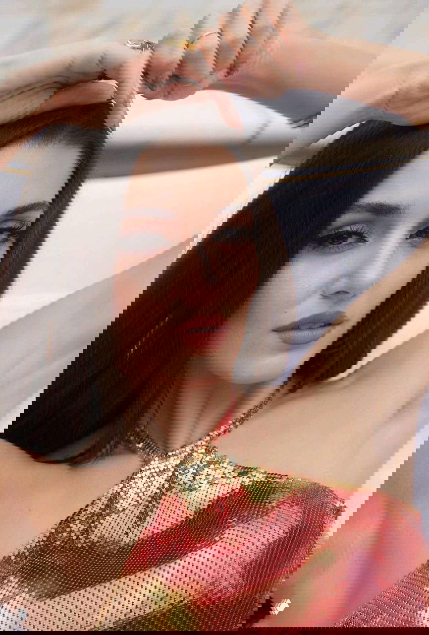 Крупный план лица женщины с минималистичным макияжем и золотистым топом, расслабленная поза и акцент на украшениях.