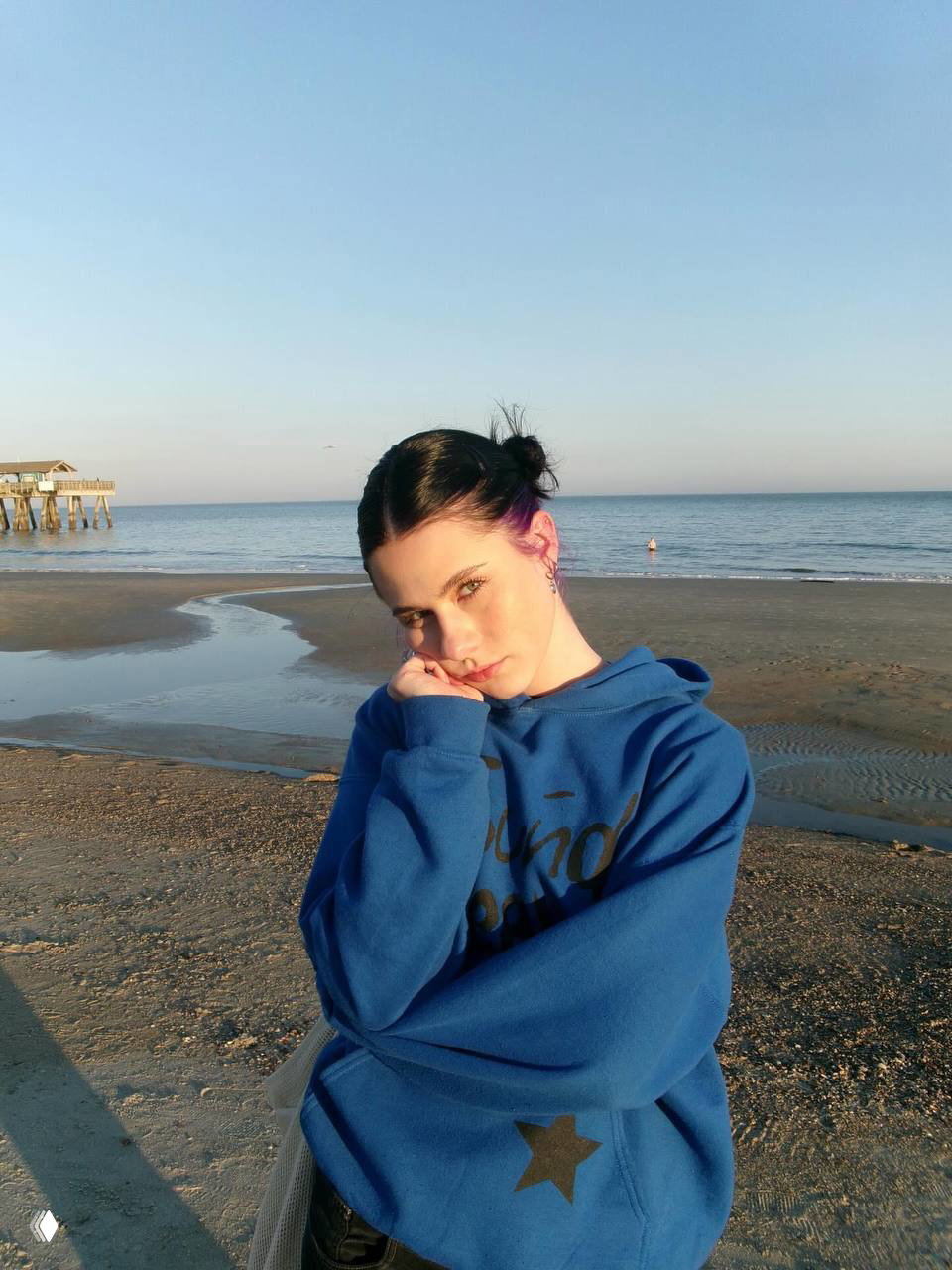Девушка в синей толстовке стоит на берегу моря; спокойный пляж, ровная линия горизонта и мягкий природный свет, задумчивый образ.