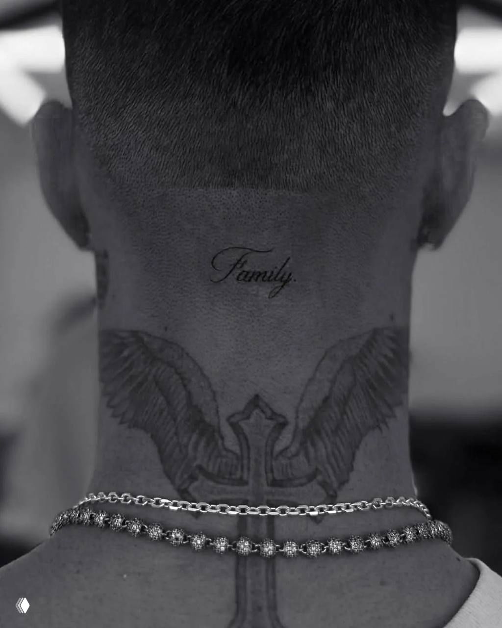 Крупный план задней части шеи молодого мужчины с аккуратной татуировкой-надписью «Family», видны цепочки и контуры других татуировок вокруг.