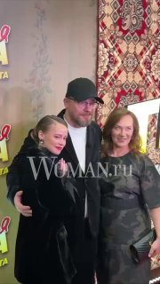 Трое людей позируют на фотозоне с узорчатым фоном и логотипом Woman.ru: мужчина в кепке и две женщины в тёмной одежде.