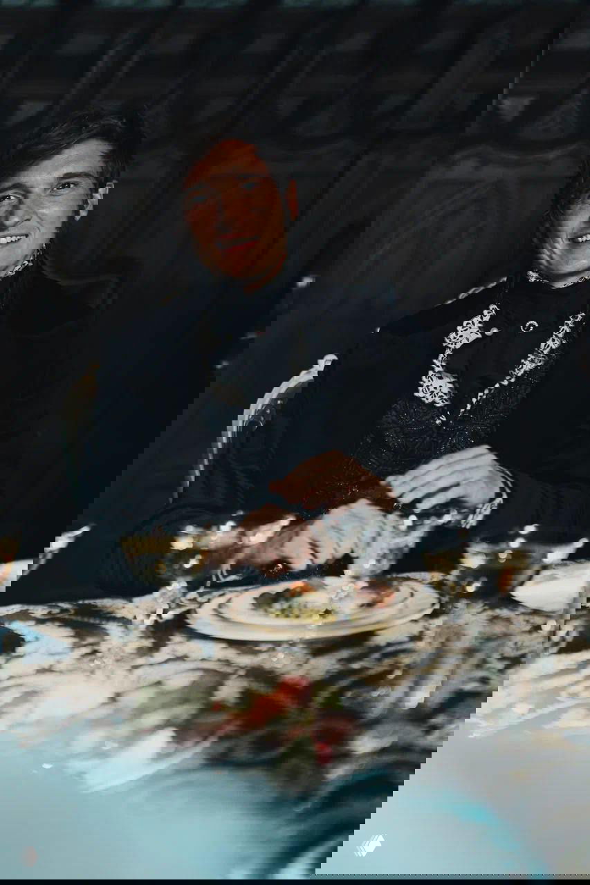 Матвей Лыков улыбается за праздничным столом в сценическом костюме XIX века; сервировка, бокалы и атмосфера зала видны на кадре со съёмок.