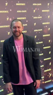 Денис Шведов в тёмном пиджаке и розовой футболке стоит на красной дорожке премьеры сериала «Чудо», на фоне виден брендовый баннер Woman.ru.
