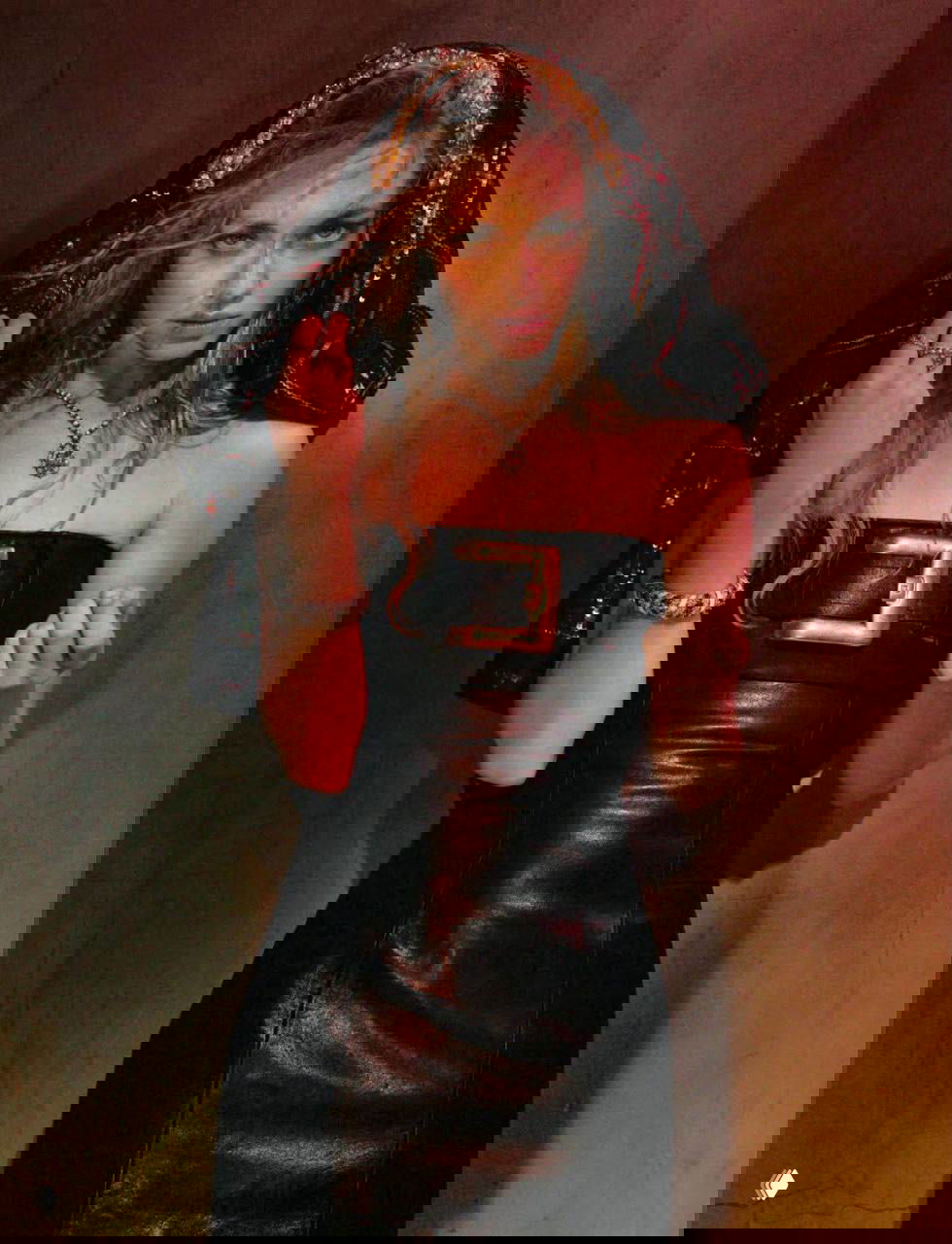 Студийный кадр в тёмном свете: женщина в кожаном платье и с украшениями стоит у стены, драматическая поза и театральное освещение.