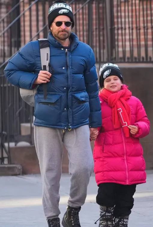 Мужчина в зимней куртке гуляет с девочкой, оба одеты тепло; городской фон с кирпичными фасадами и прохожими позади.