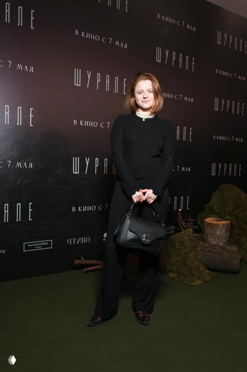 Катрин Асси в лаконичном чёрном костюме с сумкой на фоне баннера премьеры «Шурале», кадр с московского мероприятия и декораций.