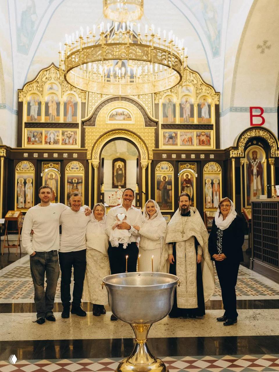 Групповое фото семьи и священника у иконостаса в храме: все участники церемонии стоят рядом с младенцем и крестильной купелью.