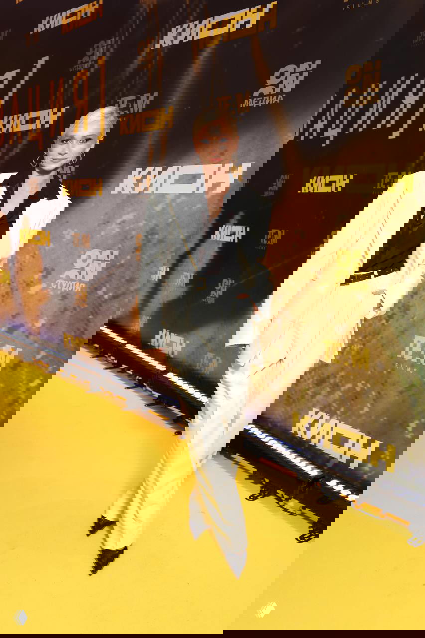 Женщина в светлом костюме и чёрной блузе на жёлтой ковровой дорожке у фона с логотипом фильма — стильный монохромный лук.