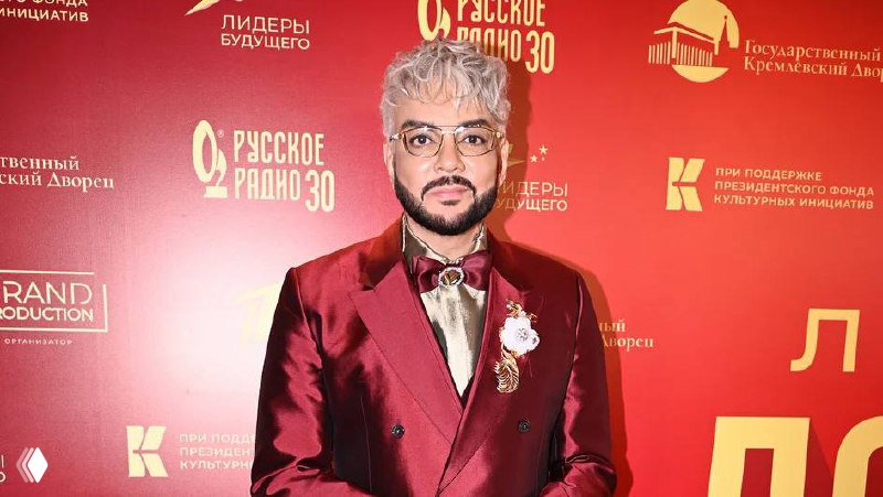 Мужчина в бордовом смокинге на красной фотозоне с логотипами мероприятий; крупный план костюма, броши и аксессуаров на лацкане.