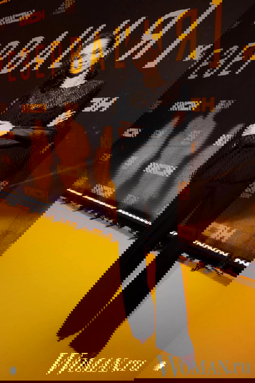 Гостья на жёлтой ковровой дорожке премьеры фильма «Резервация» в чёрном наряде с декоративными элементами, позирует у брендволла.