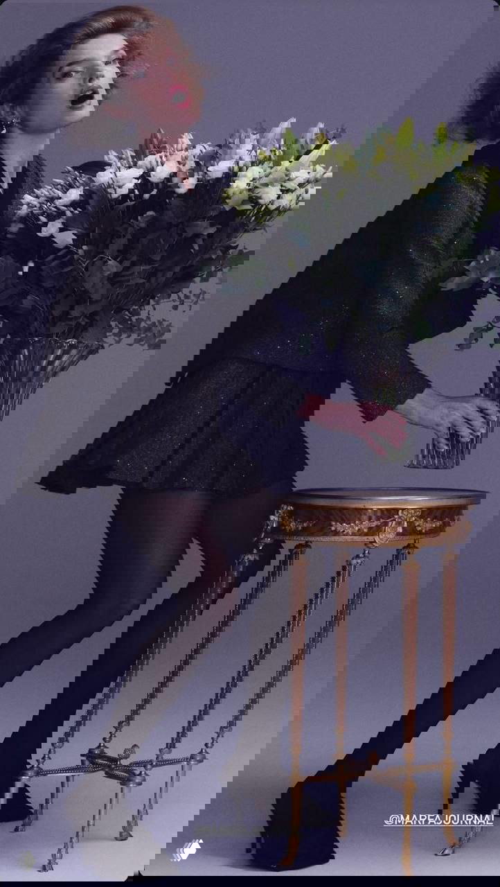 Фотоснимок в студии: женщина в чёрном костюме и мини-юбке держит большой букет белых цветов рядом с антикварным столиком.