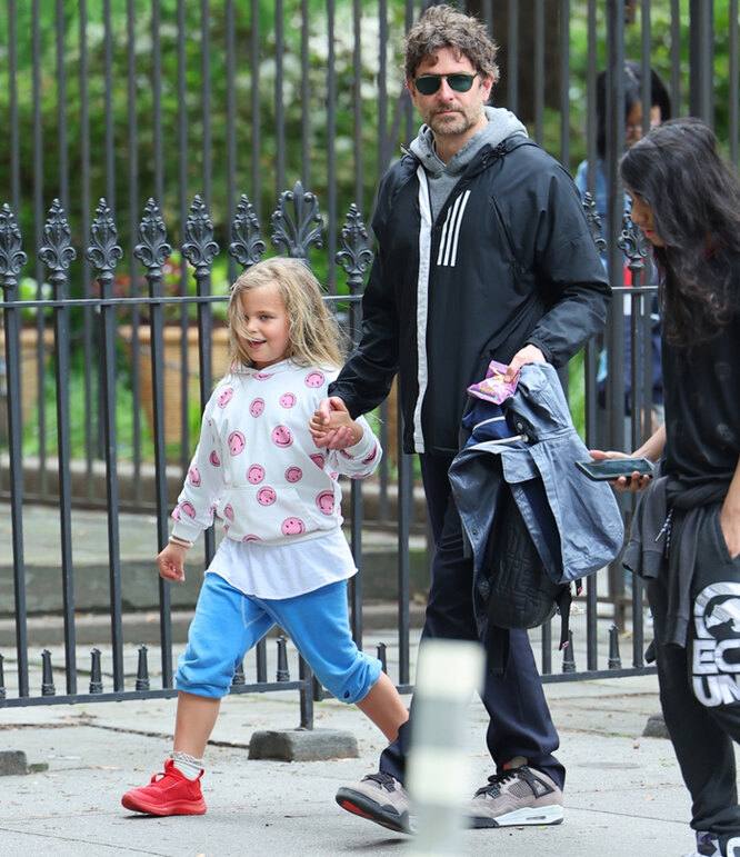 Мужчина в тёмной куртке идёт по парку, держит за руку улыбающуюся девочку; зелёные ограждения и дорожки видны на заднем фоне.