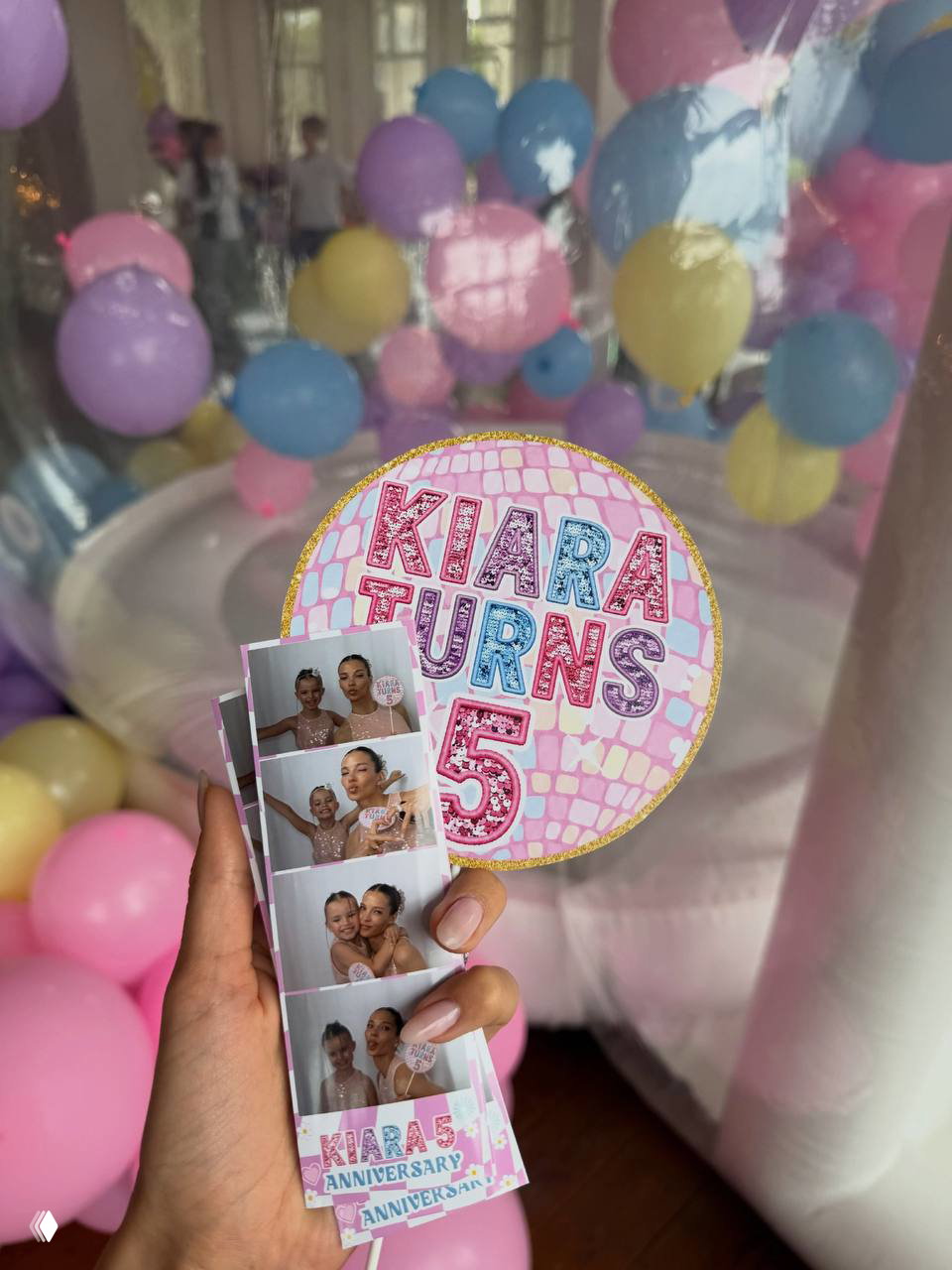Крупный план: рука держит полоску фотобута и круглый значок с надписью KIARA TURNS 5 на фоне пастельных шаров и праздничной атрибутики.