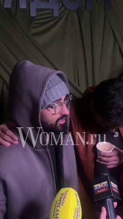 Мужчина в худи и очках стоит на фоне зелёного баннера презентации, посетители держат напитки; кадр из зоны встречи гостей и прессы.