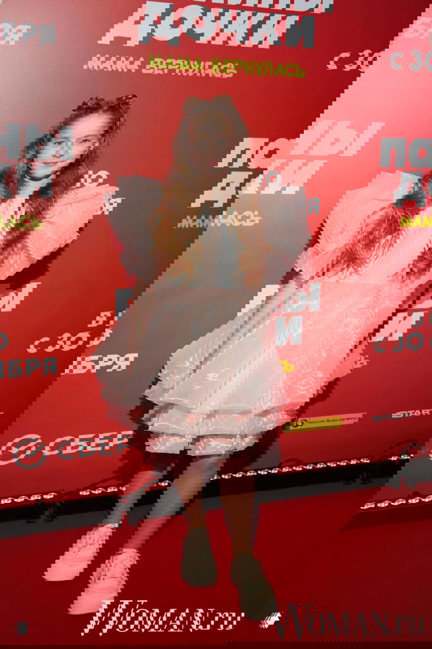 Девочка в розовом пышном платье позирует на красной дорожке у баннера премьеры «Папины дочки. Мама вернулась», на фото присутствует логотип Woman.ru.