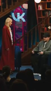 Фрагмент студийной сцены: женщина в длинном плаще стоит перед сидящим ведущим, на фоне яркая декорация и зрительный зал ток‑шоу.