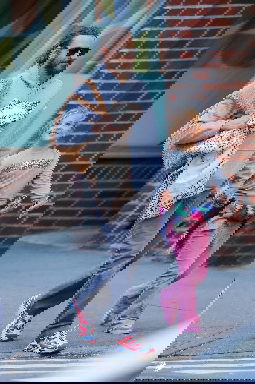 Мужчина в светлой толстовке и солнечных очках идёт по улице, держа за руку девочку; городской пейзаж и прохожие видны позади.