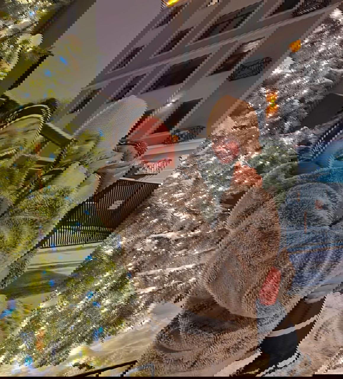 Даниил Наумов смеётся и держит ребёнка на руках возле украшенной гирляндами ёлки на заснечной улице вечером — тёплый семейный кадр.