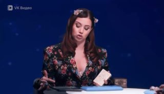 Женщина-ведущая в цветочном платье сидит за столом на телеэфире, демонстрирует жесты и держит карточку перед собой.
