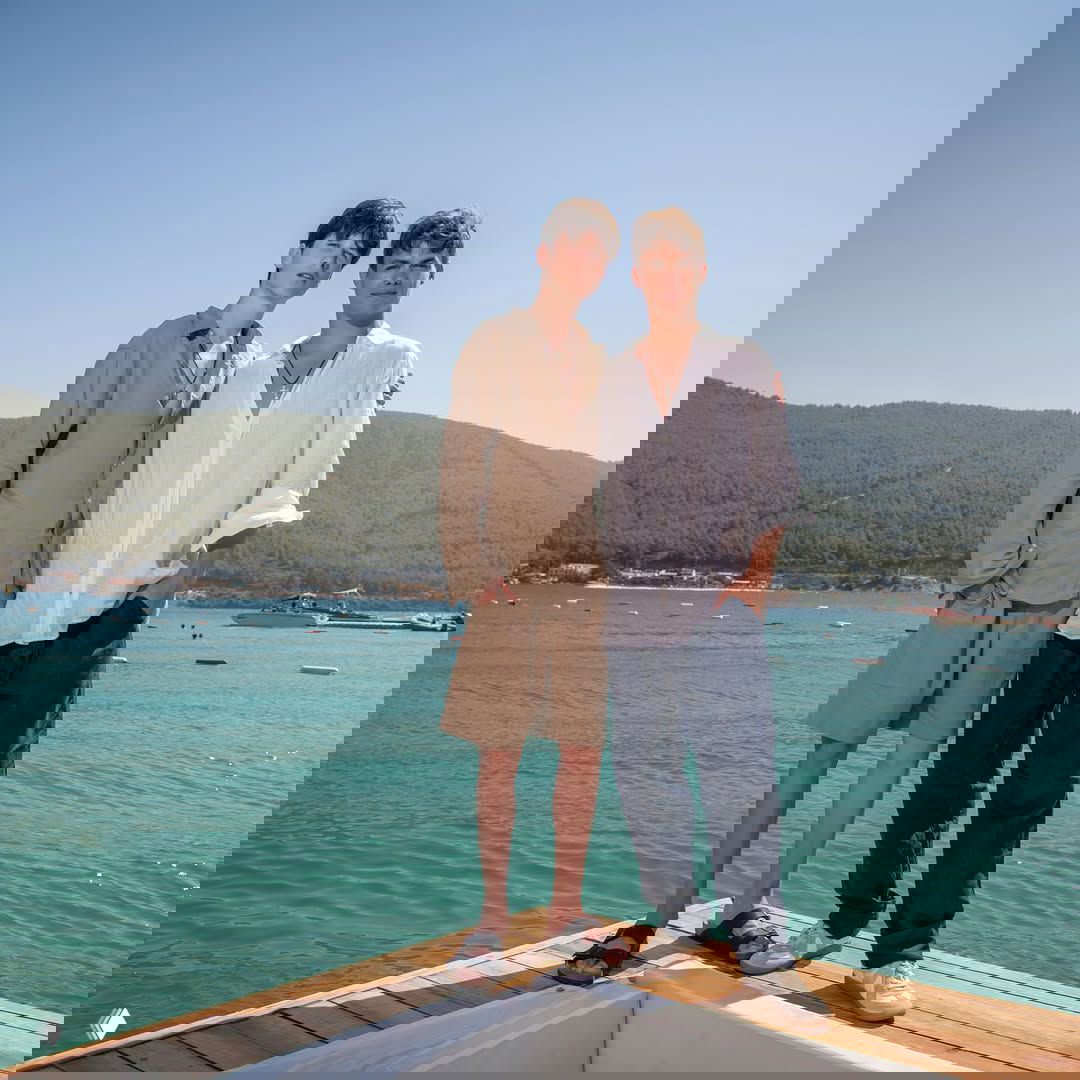 Два юноши стоят на пирсе у бирюзового моря и холмистого побережья на заднем плане, летняя одежда и морской пейзаж.