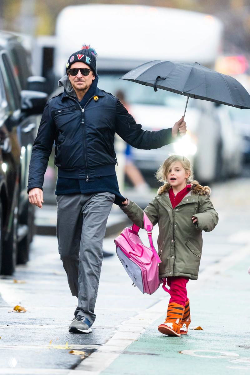 Мужчина поднимает зонт и держит девочку за руку во время дождливой прогулки; мокрый тротуар и транспорт на заднем плане.