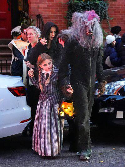 Уличный снимок: мужчина в костюме и девочка в карнавальном платье идут по улице во время мероприятия, вокруг люди и припаркованные автомобили.