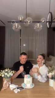 Пара в гостиной улыбается у праздничного торта и цветов — кадр из видео, где супруги узнают пол первенца; ожидается девочка.