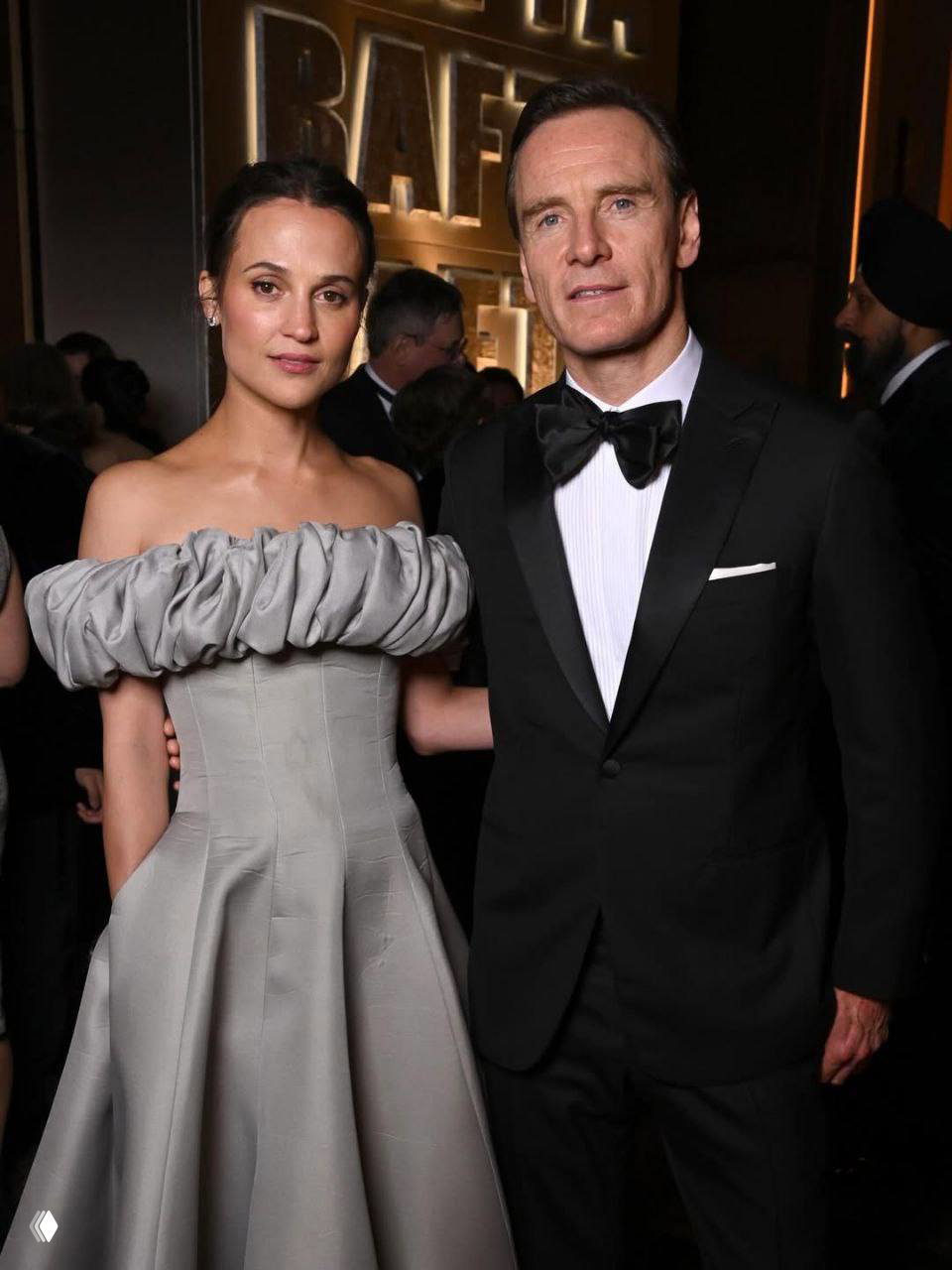 Крупный план: Алисия Викандер и Майкл Фассбендер в вечерних нарядах позируют на фоне логотипов BAFTA на мероприятии в Лондоне.